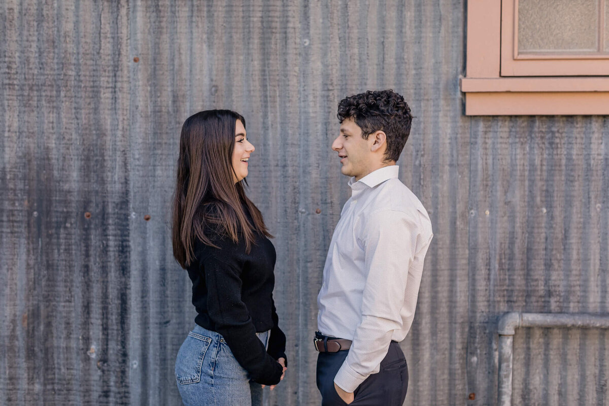 A brother and sister with dark brown hair standing facing each other and laughing in front of a grey, metal wall.