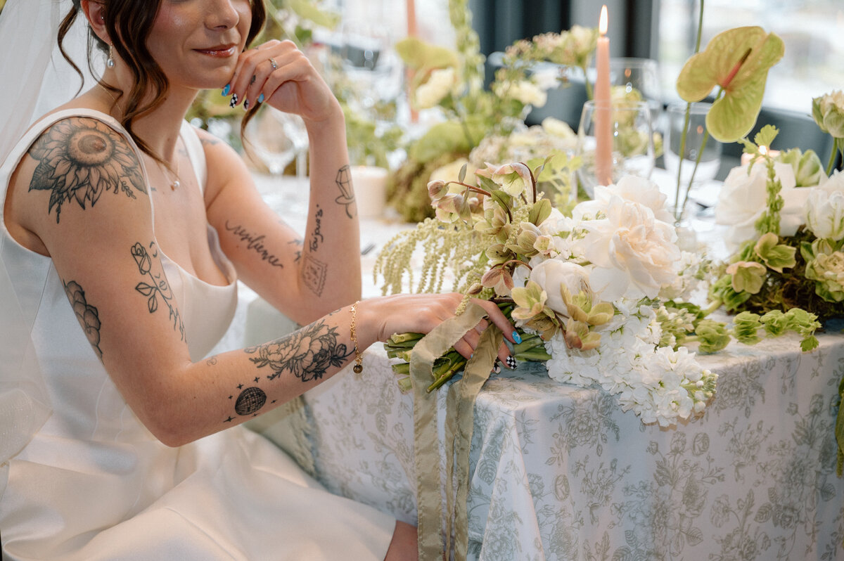 Bride with tattoos sits at floral reception table in editorial wedding photo.