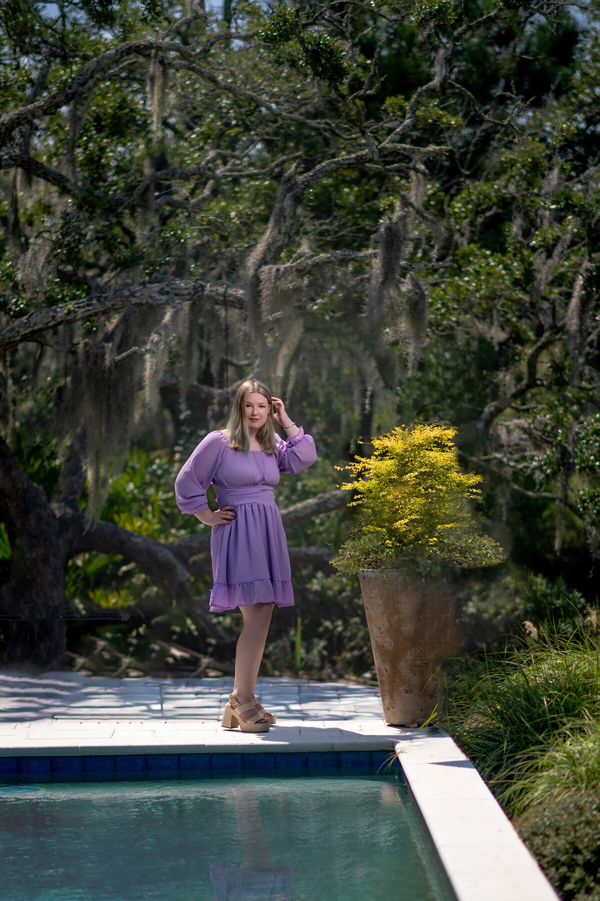 A woman in a purple dress stands by a pool under oak trees with a hand on her hip