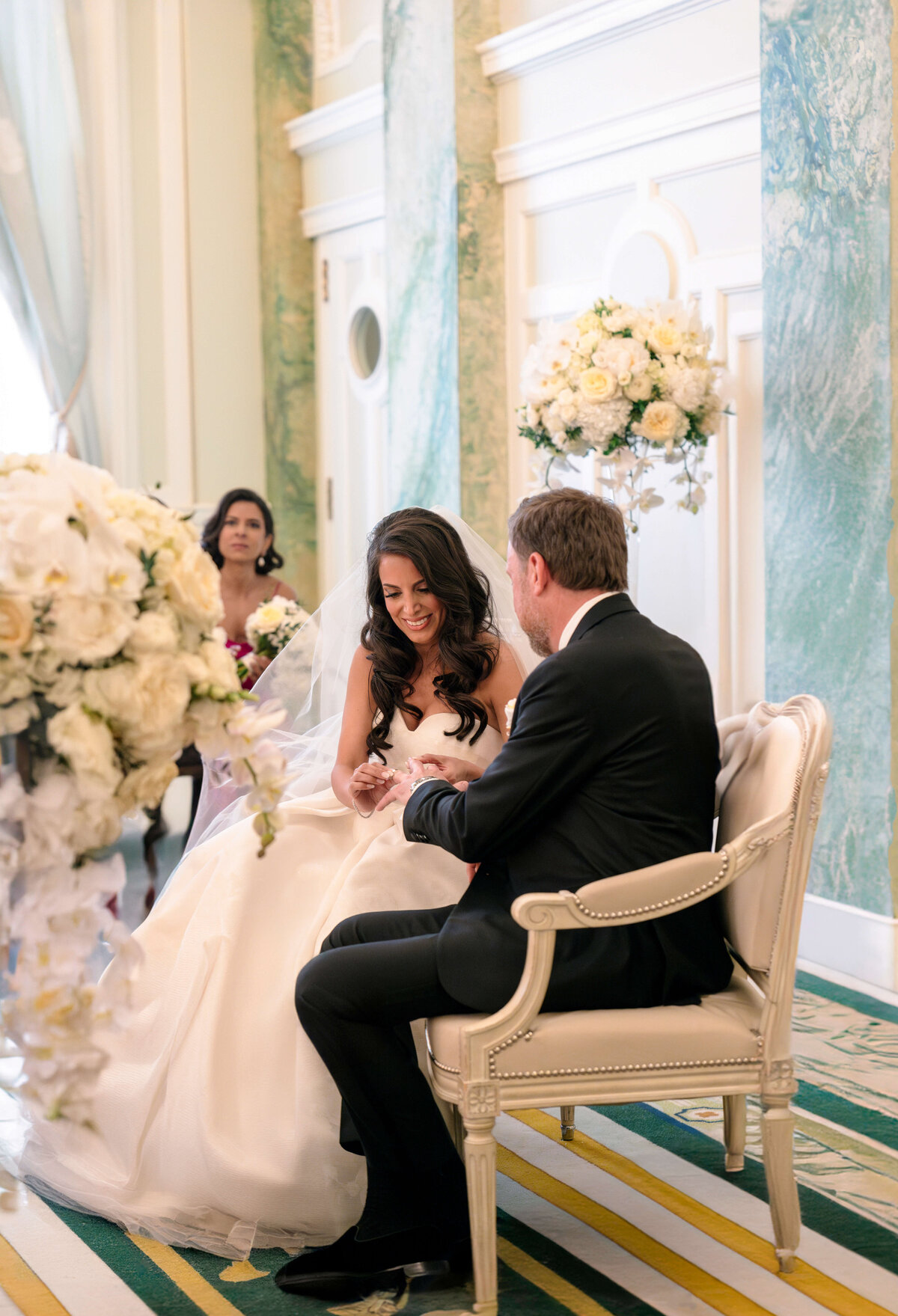 A bride and groom sit smiling and holding hands in a beautifully decorated room with green accents and large floral arrangements. A bridesmaid is visible in the background. The atmosphere is elegant and joyful.