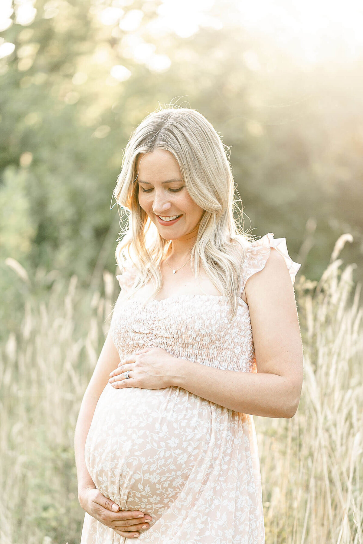 Mama holding her baby bump and looking down and smiling. She is holding her belly and wearing a beige colored floral dress and standing in a field of tall golden grasses at sunset