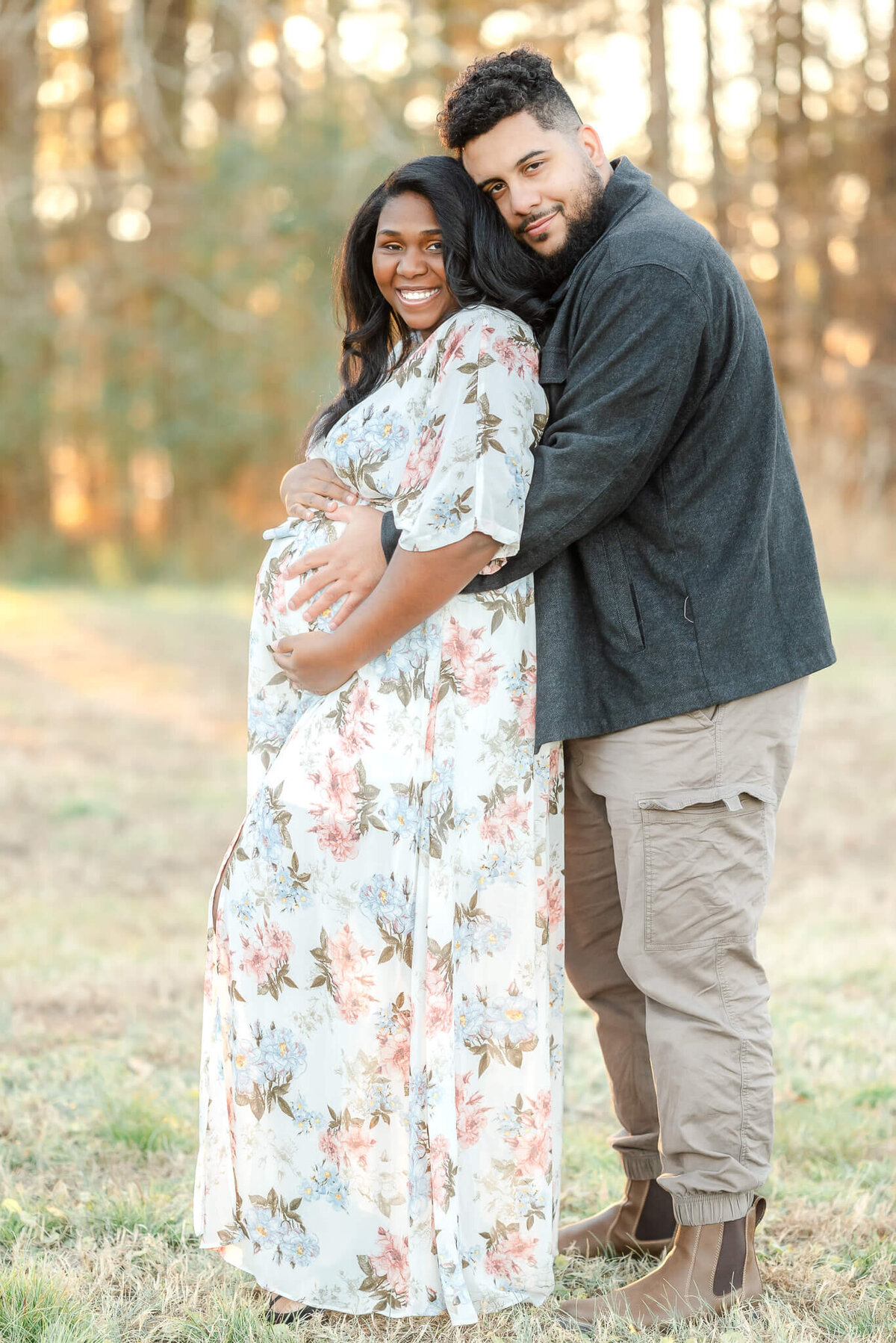 A pregnant couple snuggles close, with dad behind mom. Mom shines in a white floral dress during her Chesapeake maternity photography session.