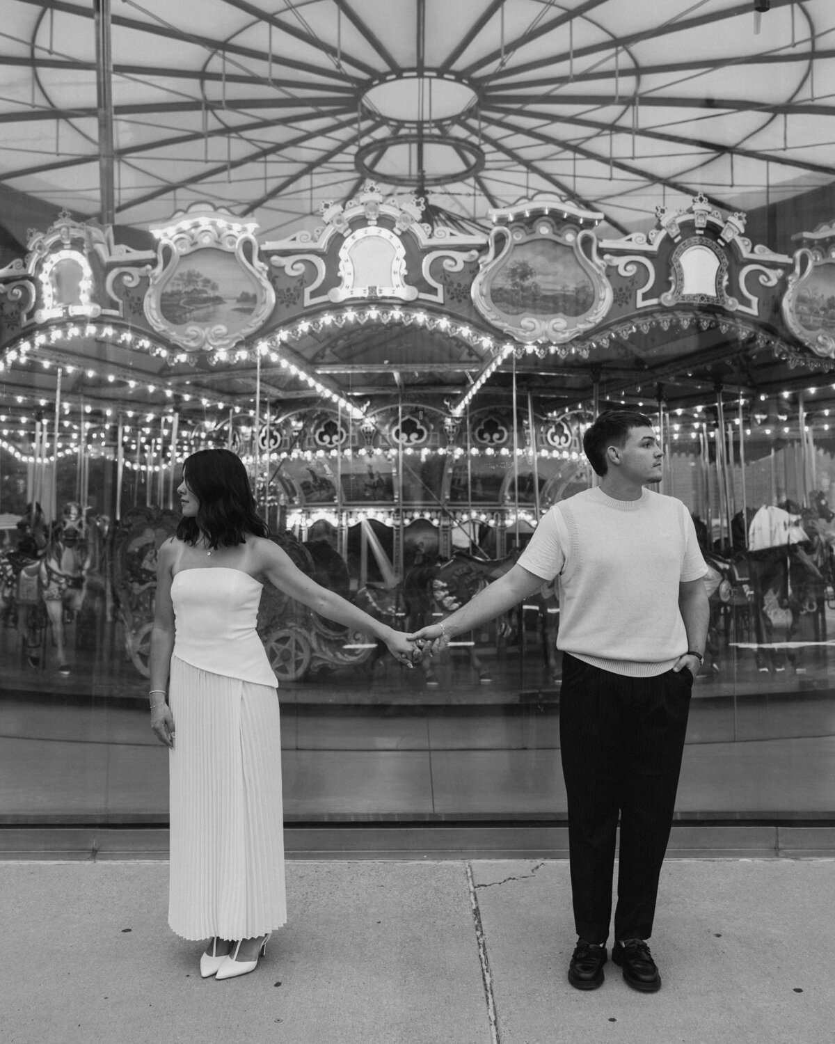 A couple stands hand in hand in front of a beautifully lit carousel, dressed elegantly amidst a nostalgic park setting.