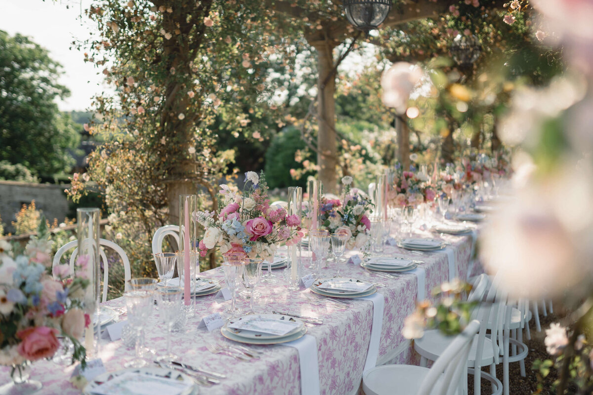 long wedding dining table with pink and white patterned linen set under a rose arbour in euridge manor’s gardens for a romantic cotswold wedding