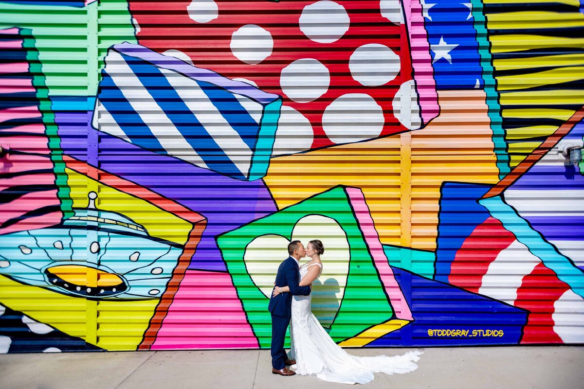 A wedding couple kissing in front of a large, colorful mural.