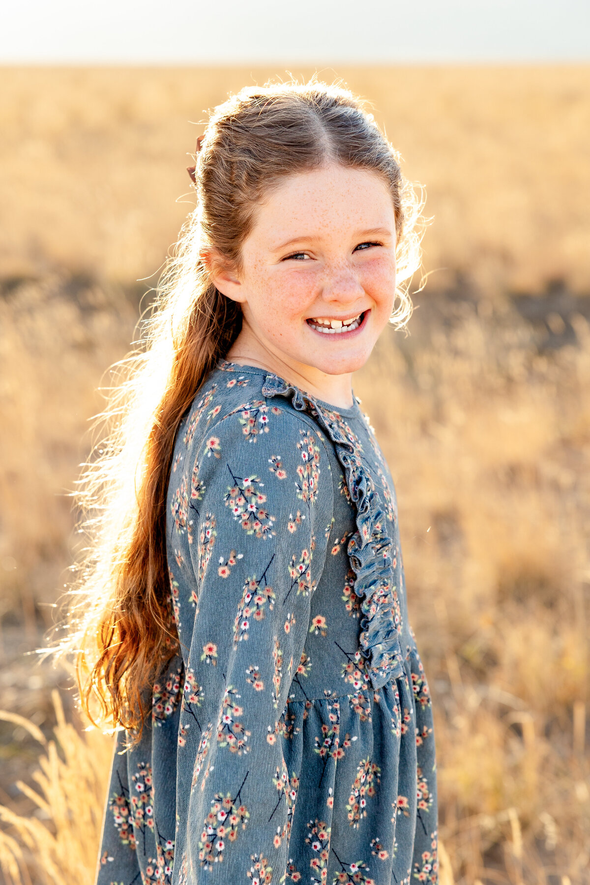 A freckle-faced girl smiles brightly as the sunlight highlights her long, wavy hair in a golden field in Eagle Mountain, Utah.