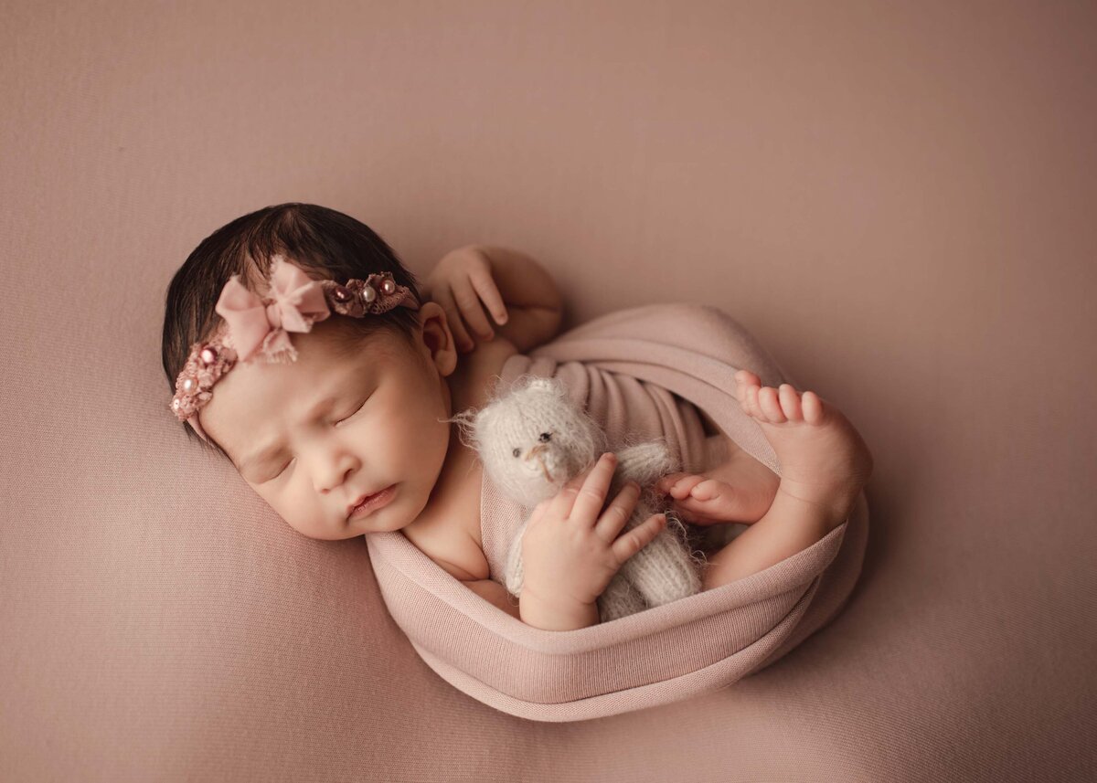 Styled newborn photo. Baby sleeping on back  wrapped in pink holding a tiny teddy bear