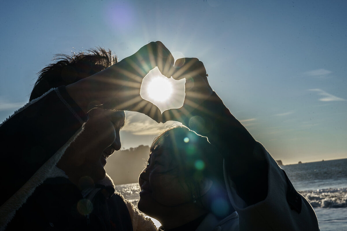 couple forming a heart shape with their hands and the sunset shines through the heart