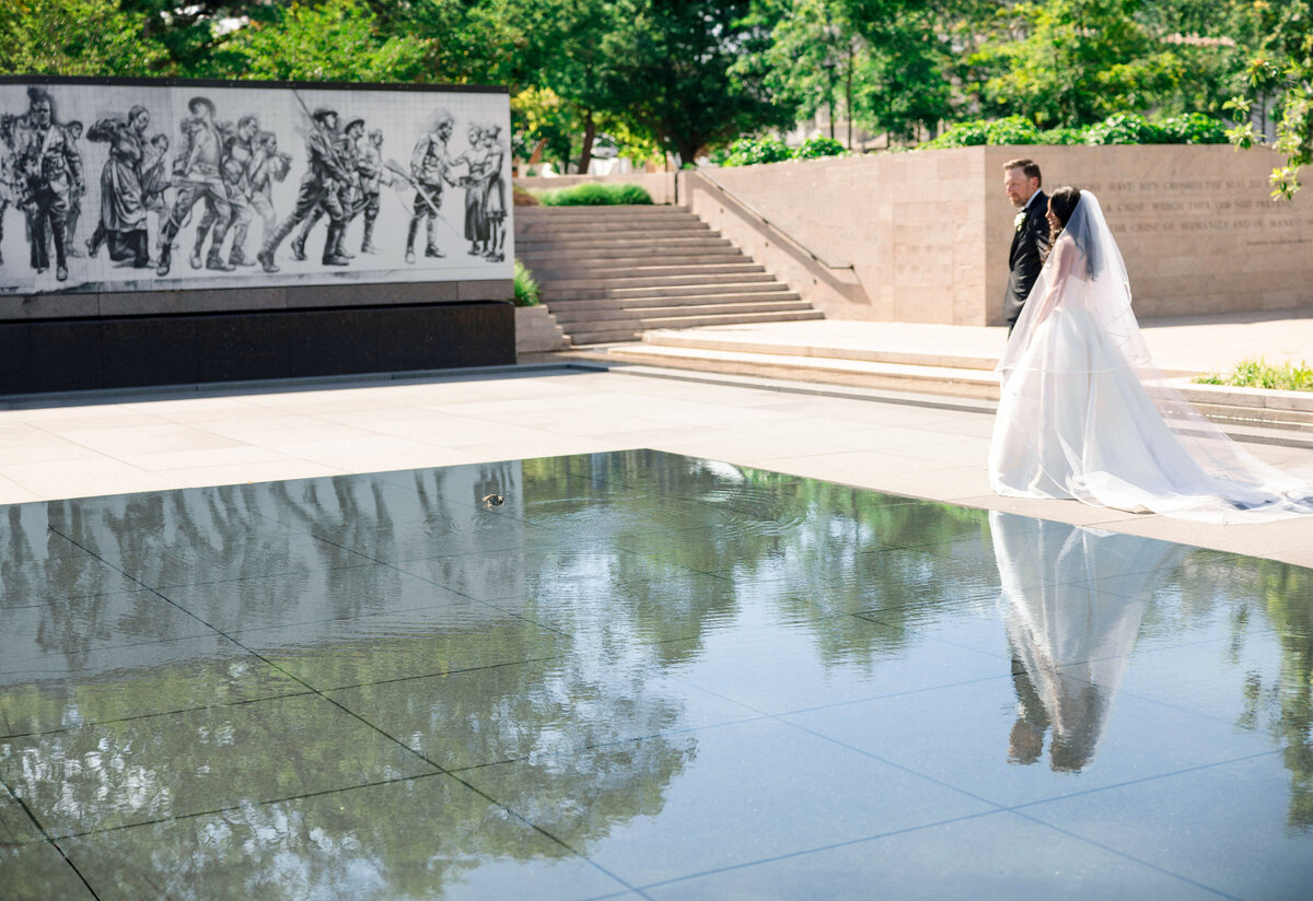 A bride and groom walk beside a reflective pool. Behind them is a wall with engraved historical images. The scene is surrounded by green trees and stone steps under a clear blue sky.