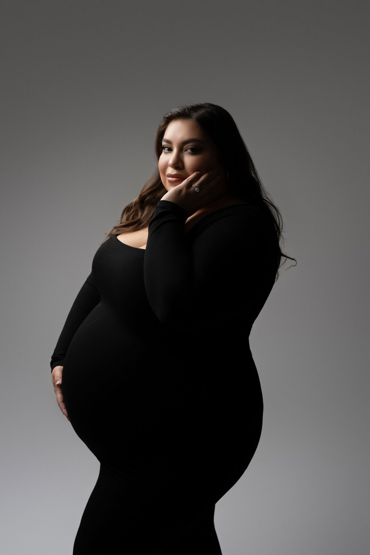 Confident maternity portrait of an expecting mother in a sleek black gown, captured with soft directional lighting in studio.