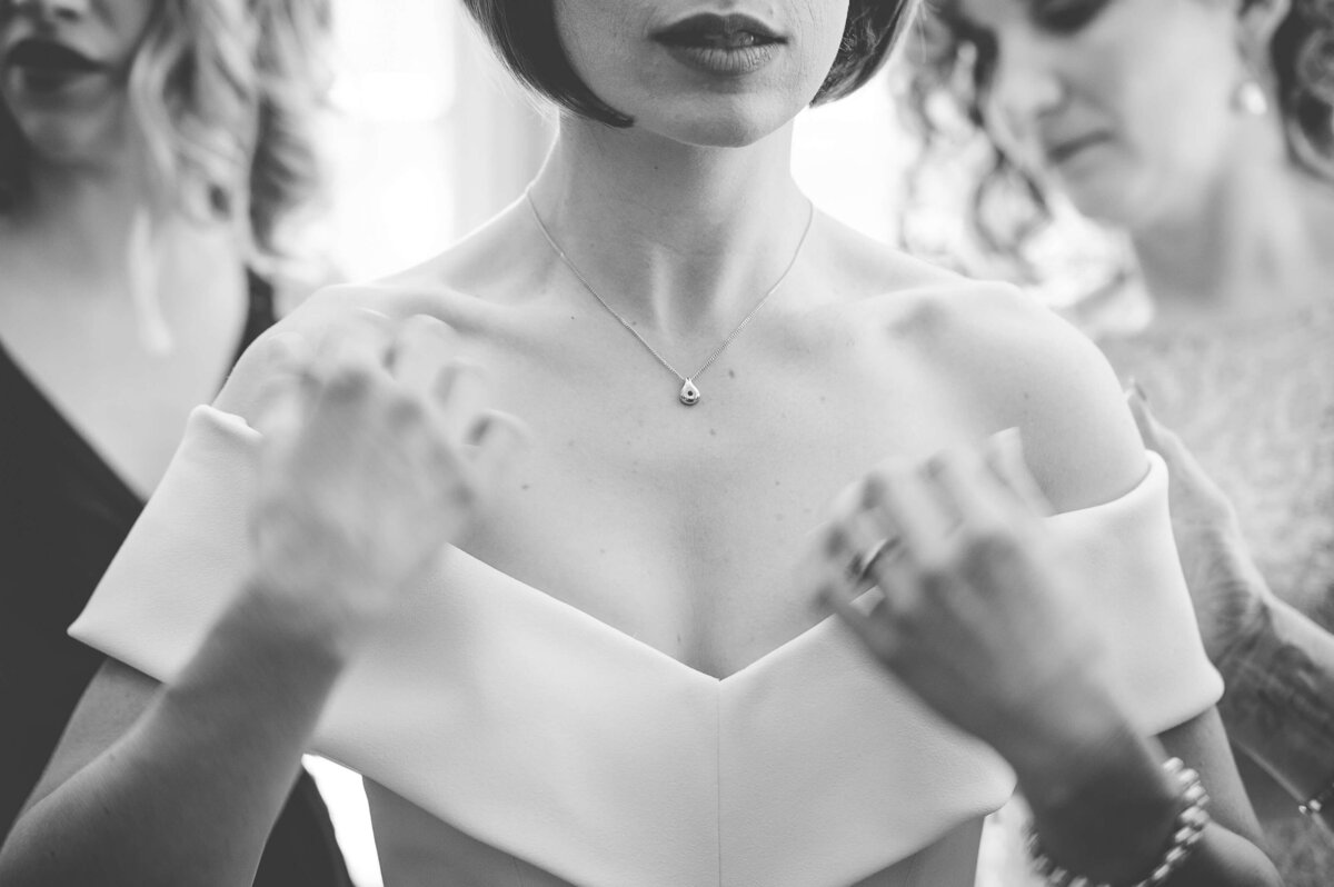 a black and white closeup photo of a bride's necklace as she gets ready with two bridesmaids behind her.  Captured by Ottawa wedding photographer JEMMAN Photography