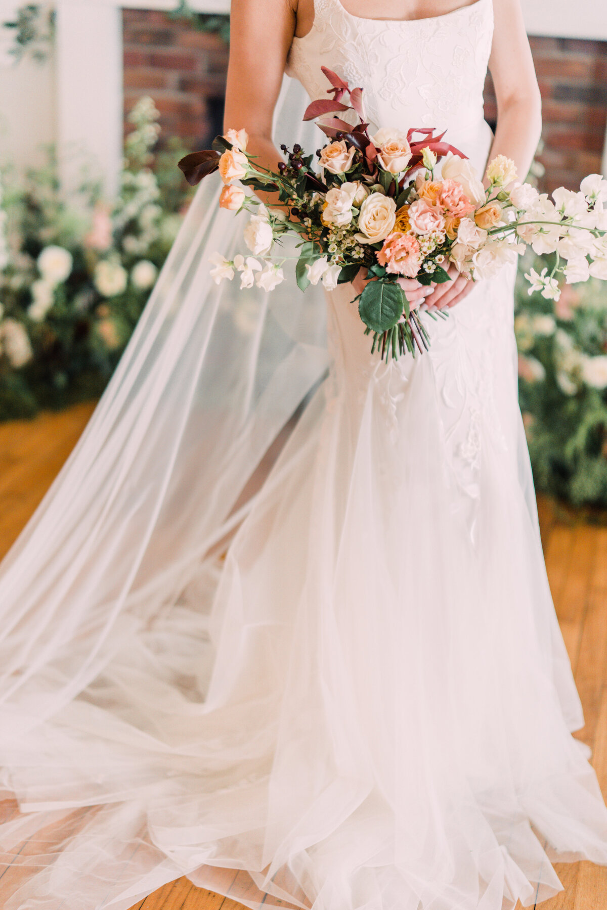 Bride wearing Alexandra Grecco Marcelle gown & Ainsley veil holding burgundy, blush, ivory, & peach bouquet at Cornman Farms