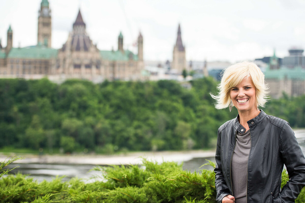 a photo of a blonde-haired woman in a black leather jacket with the Parliament buildings behind her.  Captured outdoors by Ottawa Branding Photographer JEMMAN Photographer COMMERCIAL