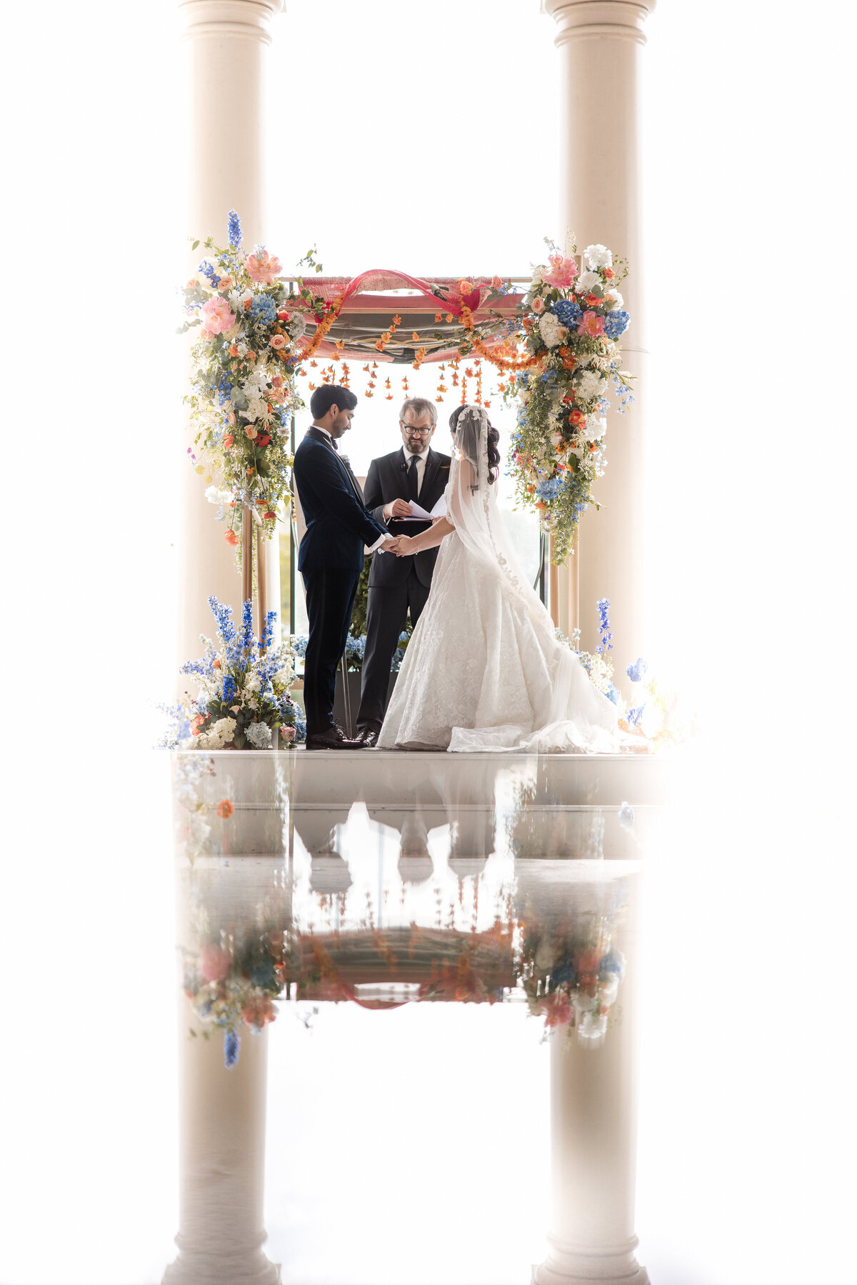 A couple holds hands beneath a floral chuppah framed by two grand columns during a wedding ceremony at Water Works in Philadelphia. The image features a symmetrical reflection of the couple, officiant, and vibrant flowers, creating a mirrored effect with soft, natural light surrounding the scene.