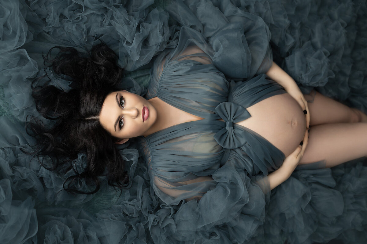 Pregnant woman lying on layers of teal tulle, hands gently resting on her belly, wearing a sheer flowing gown.