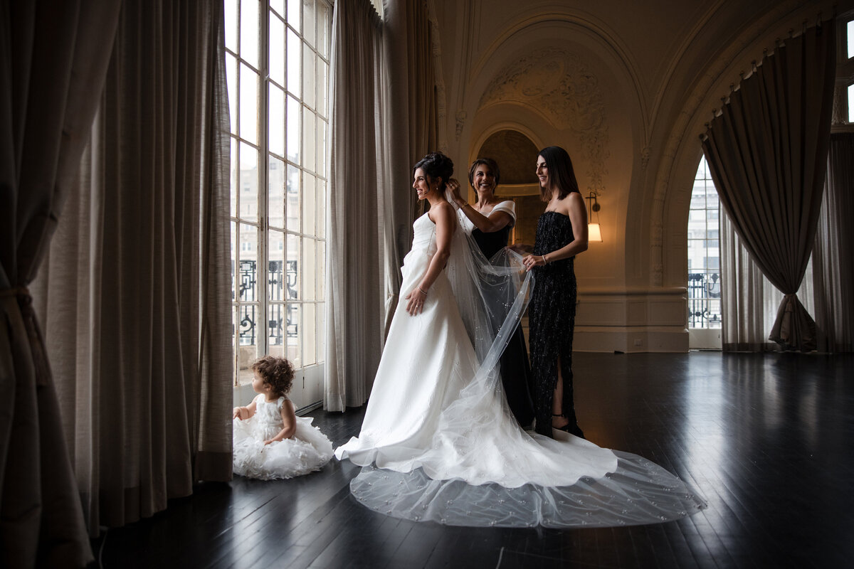 A bride prepares for her wedding day at 19, with family helping arrange a flowing veil in a softly lit room featuring tall arched windows and ornate details, while a young child in a tulle dress sits by the window—an elegant pre-ceremony moment ahead of a celebration at The Grand Bell.