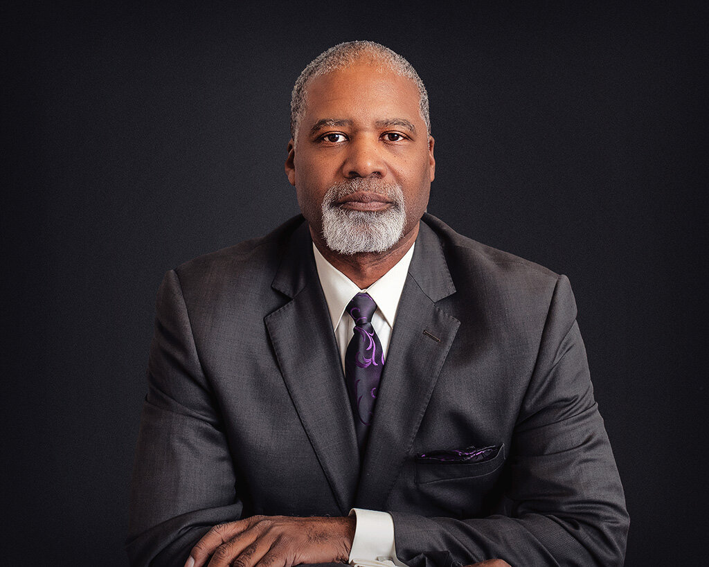 headshot of an African amercan man in a dark grey suit with a grey and purple tie, salt and pepper hair and a beard