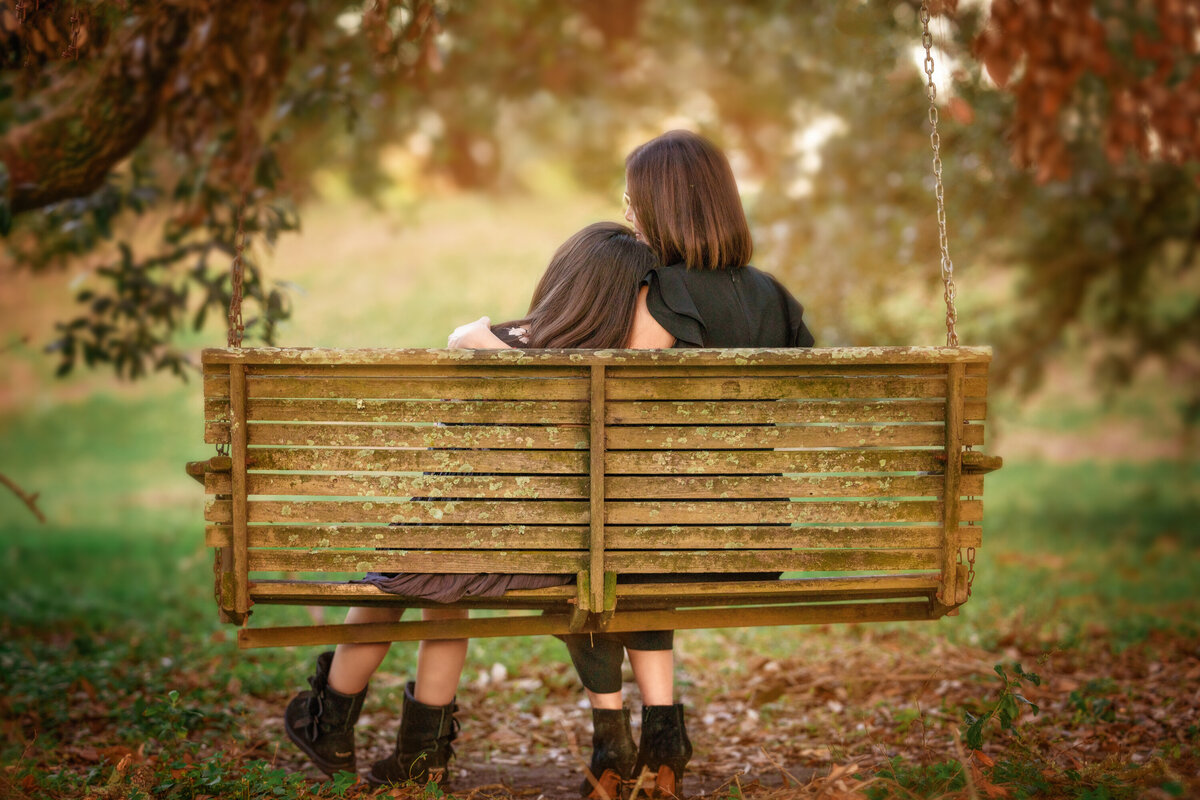 Mom and daughter, both with dark hair, and sitting on a swinging bench in a park.  The bench is wooden with slats and the trees are changing color for fall.  The daughter is resting her head on the mom's shoulder.