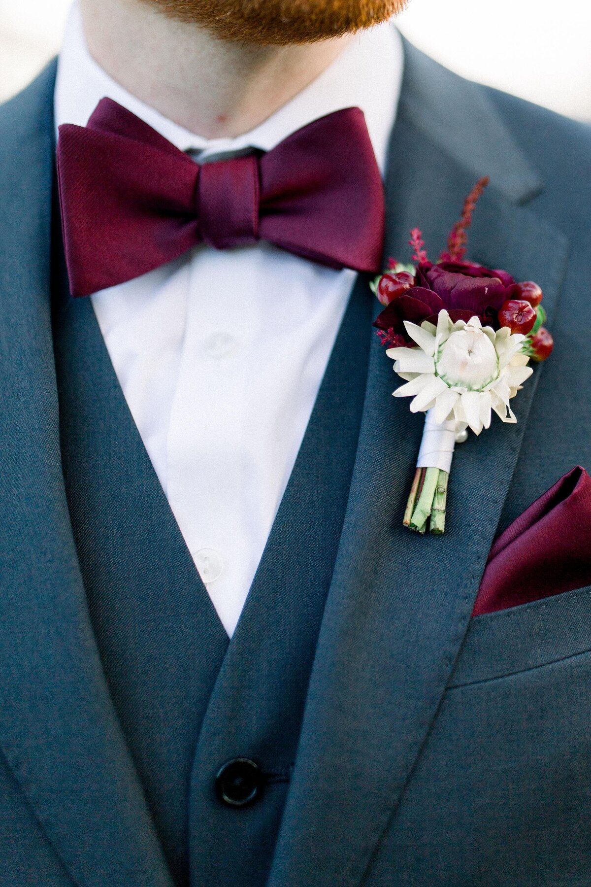 Groom wearing navy suit with burgundy bow tie featuring a white and burgundy boutonniere