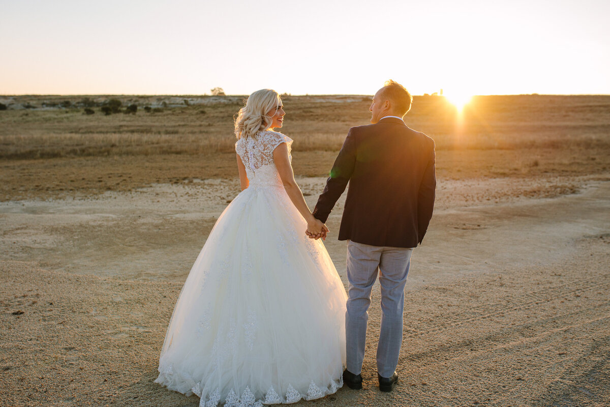 Groom & Bride walking across a flat saltpan toward the sunset, with the golden sun low on the horizon behind them.