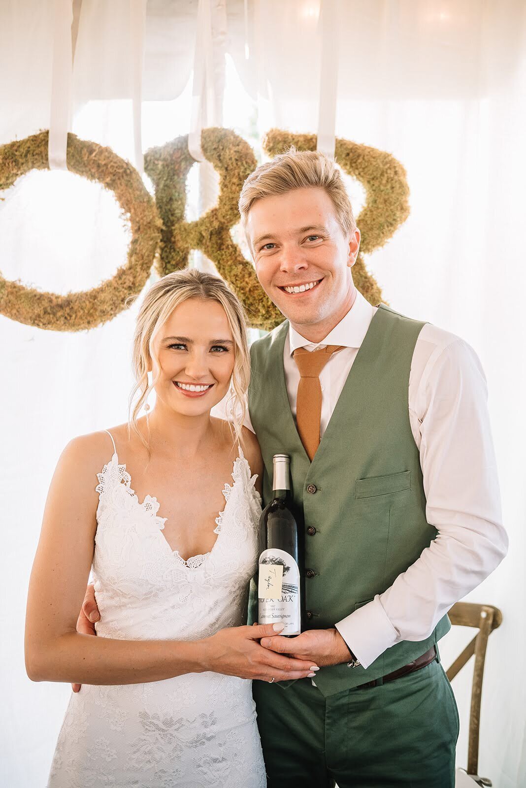 Bride and groom pose with a bottle of wine on their wedding day.