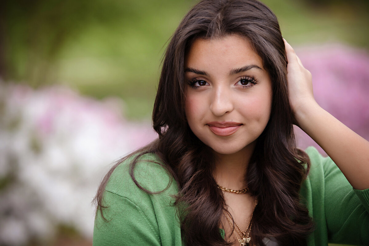 Close-up of high school senior looking at the camera in green cardigan and white tank top, framed by soft spring azaleas and natural light.