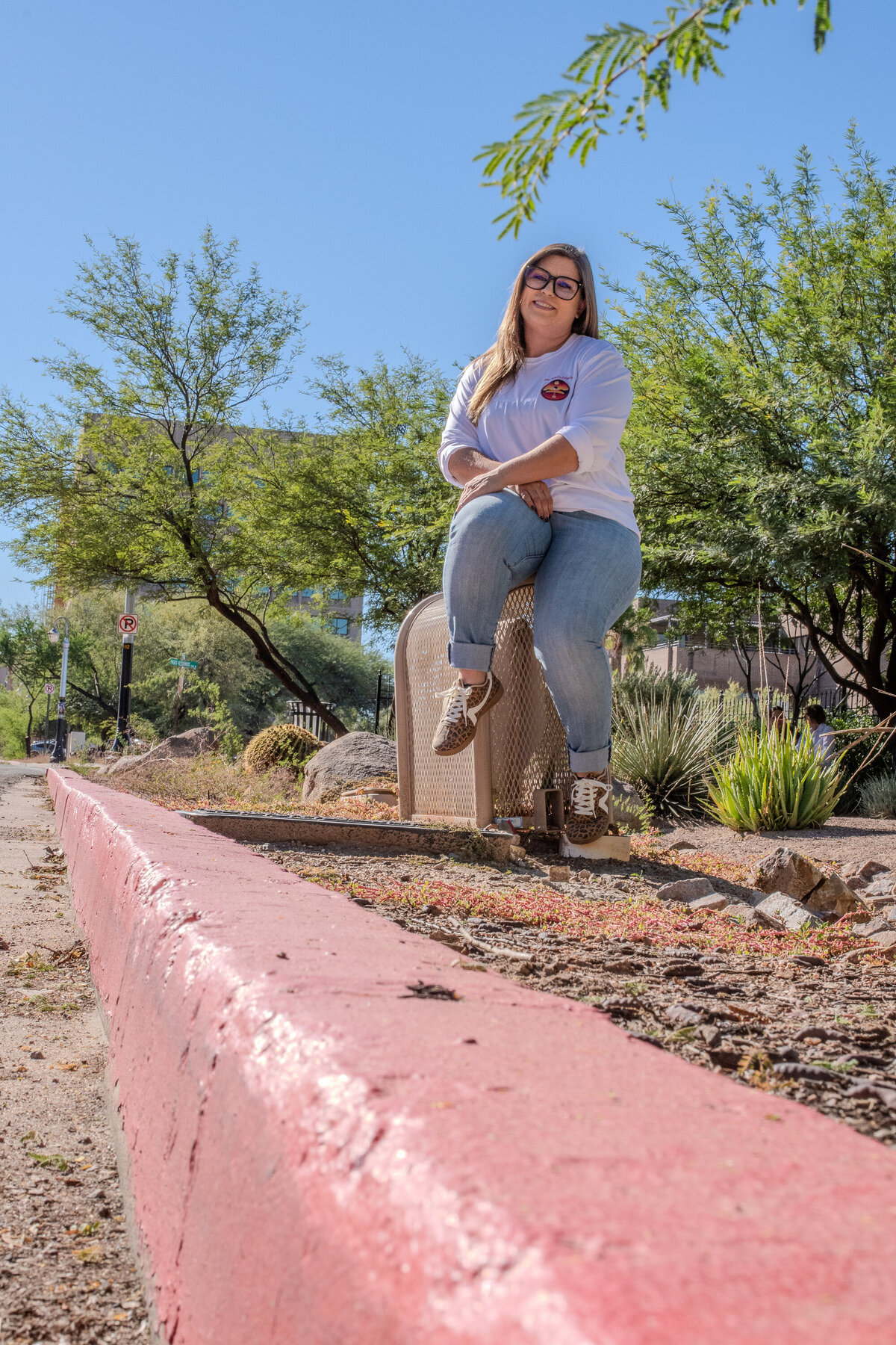 Woman sitting outdoors on a concrete post along a landscaped walkway, smiling in casual jeans and a white top, photographed by Vyrl Photo, showcasing Tucson brand photography.