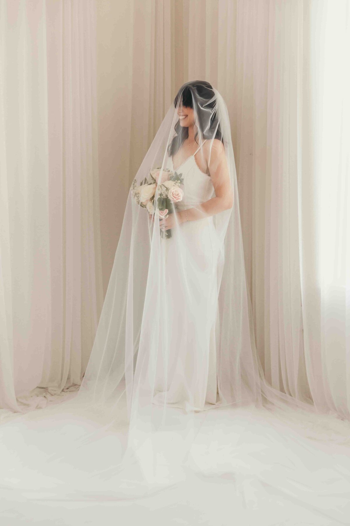 bride under veil holding bouquet in front of white curtains