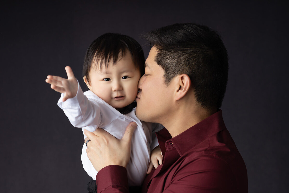 Dallas baby photographer captures a heartwarming moment as a father kisses his son dressed in a tuxedo, sharing a joyful moment.