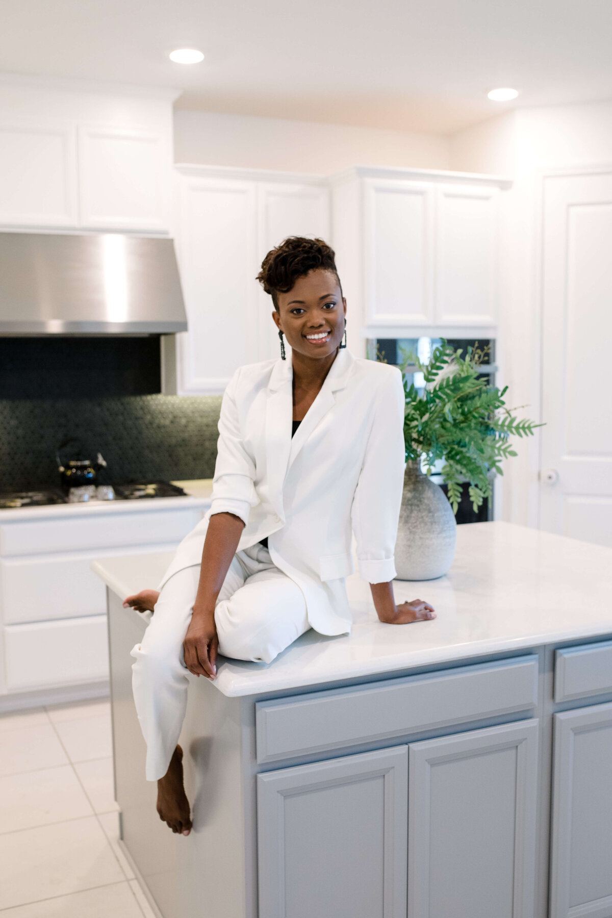 In-home brand photography with woman sitting on an island in the middle of her kitchen wearing a white blazer and smiling for the camera with her legs crossed