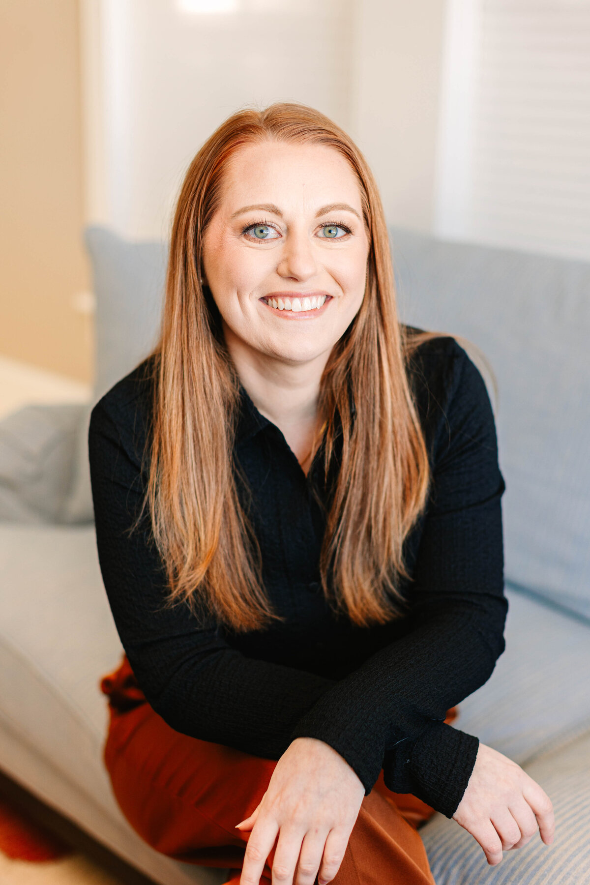 A woman with long, straight auburn hair and blue eyes sits on a light blue couch, smiling at the camera. She wears a black blouse and rust-colored pants, with her hands resting casually in front of her. The background features a neutral-toned living space.