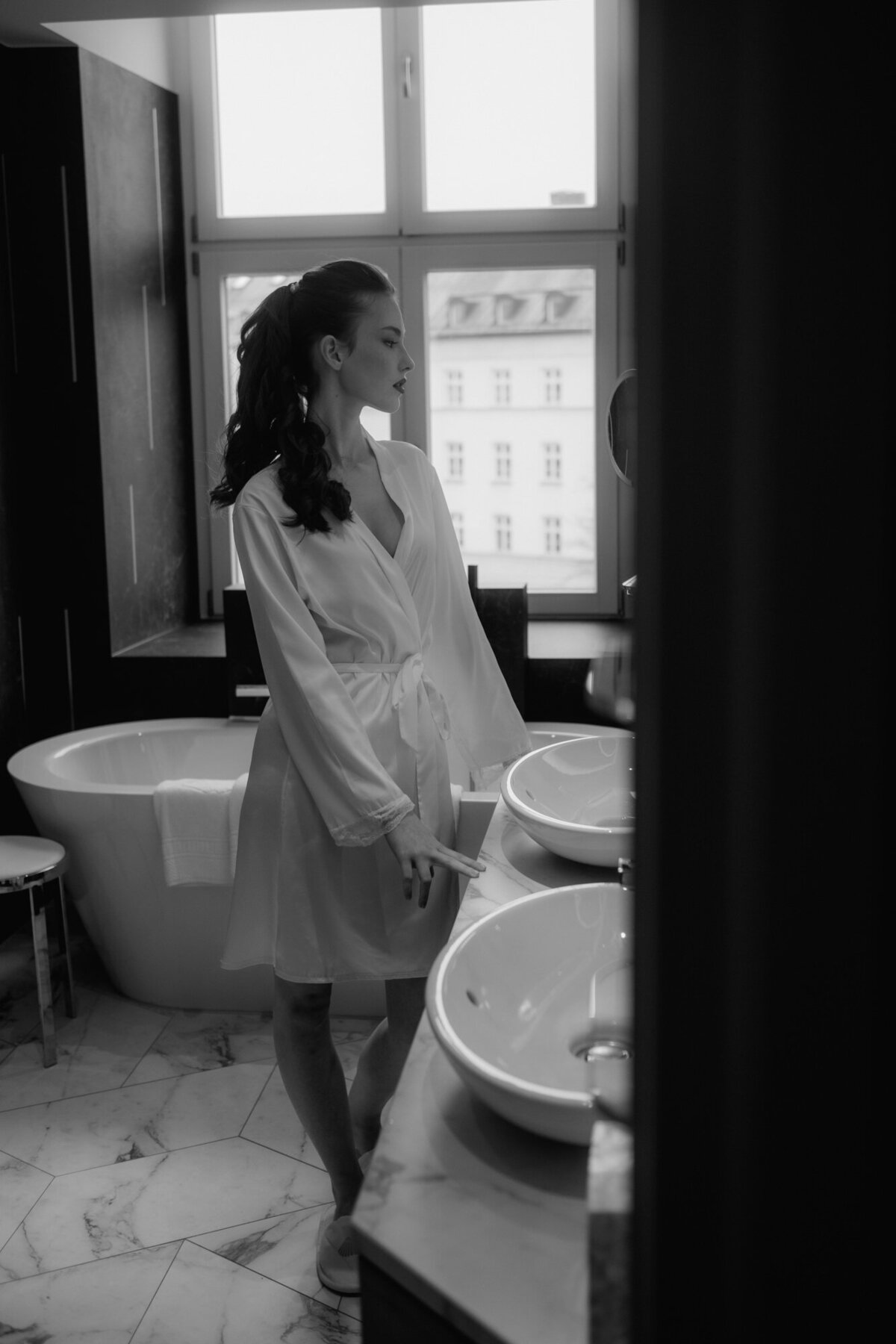 Im Badezimmer vor dem Waschbecken steht das Brautmodel im Morgenmantel mit einer Badewanne im Hintergrund.