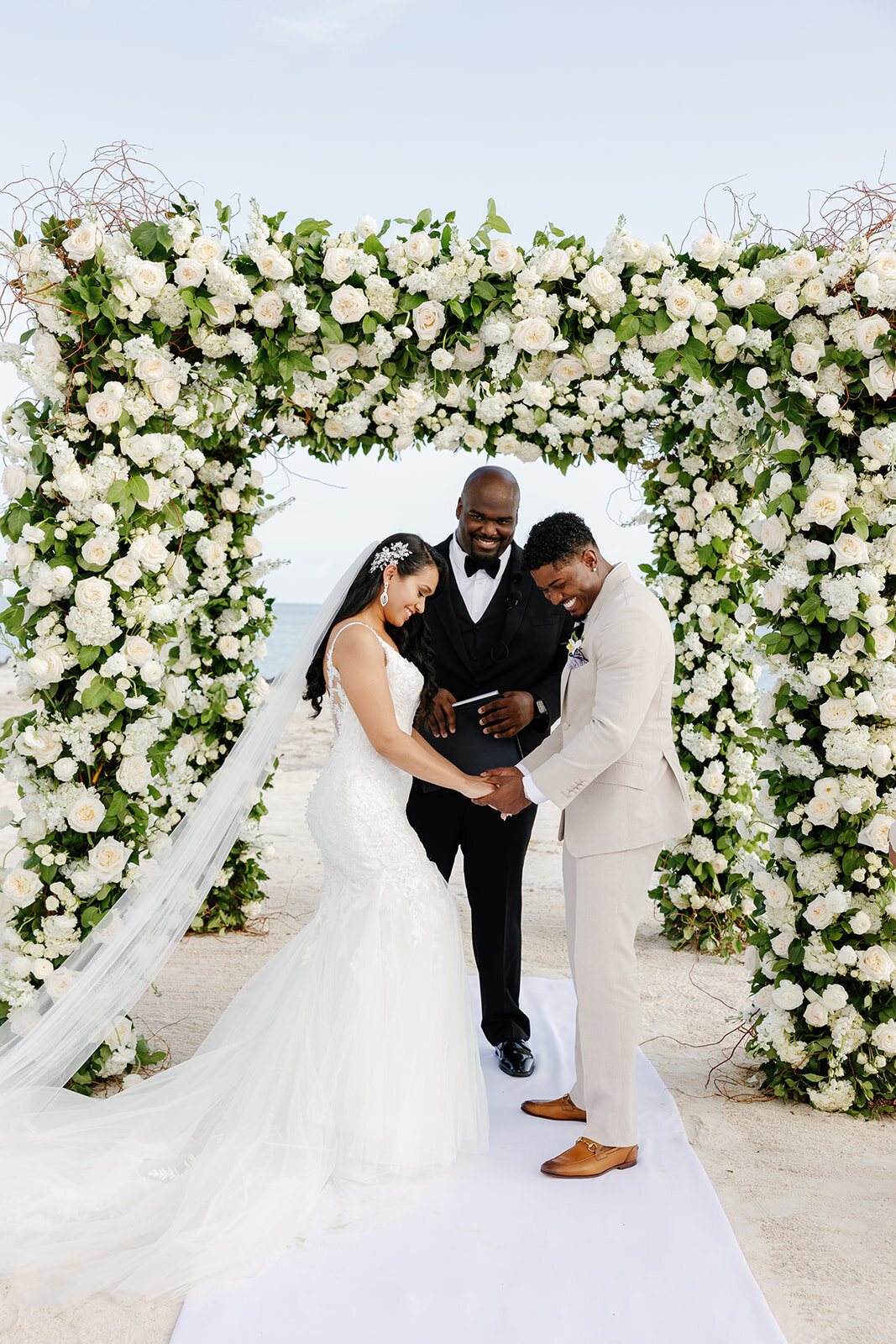 Andressa and Jonathan hold hands beneath a breathtaking floral arch, exchanging vows at their oceanfront ceremony in Table Key, Islamorada. Photography by Claudia Amalia, destination wedding photographer.