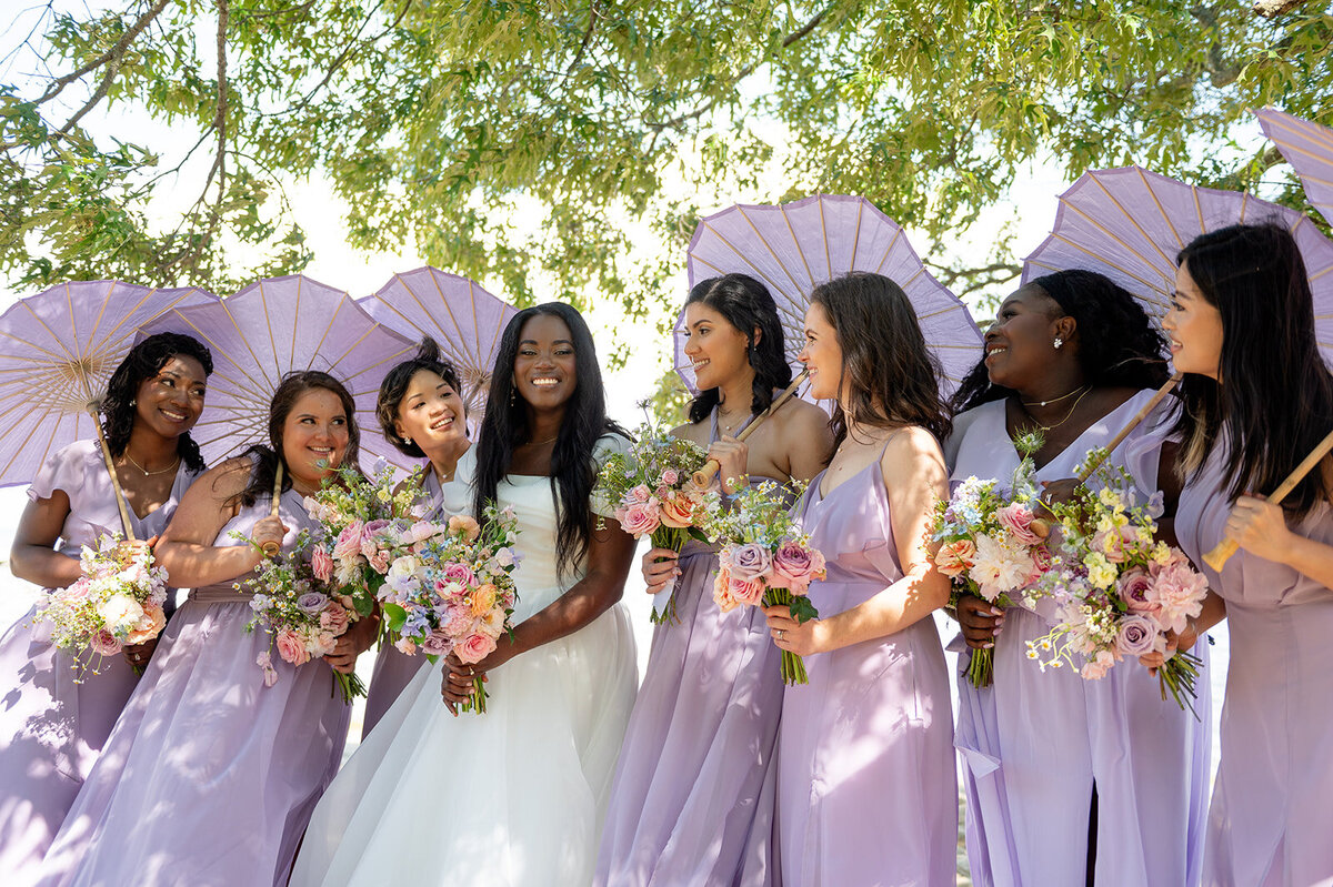 Bride and bridesmaids dressed in lavender gowns holding colorful bouquets, posing under matching lilac umbrellas., under the  trees at the Chesapeake Bay Foundation.
