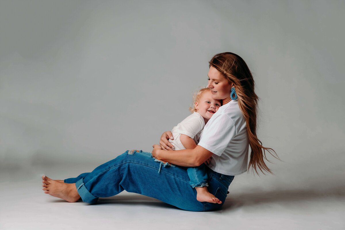 A young mom and her toddler daughter pose for a tender portrait by Mobile, AL Motherhood Photographer. The mom is seated with the child resting her head on her chest, both wearing matching jeans and shirts and barefoot. The mother looks at the camera with a warm smile, while the child enjoys a cozy moment in her lap