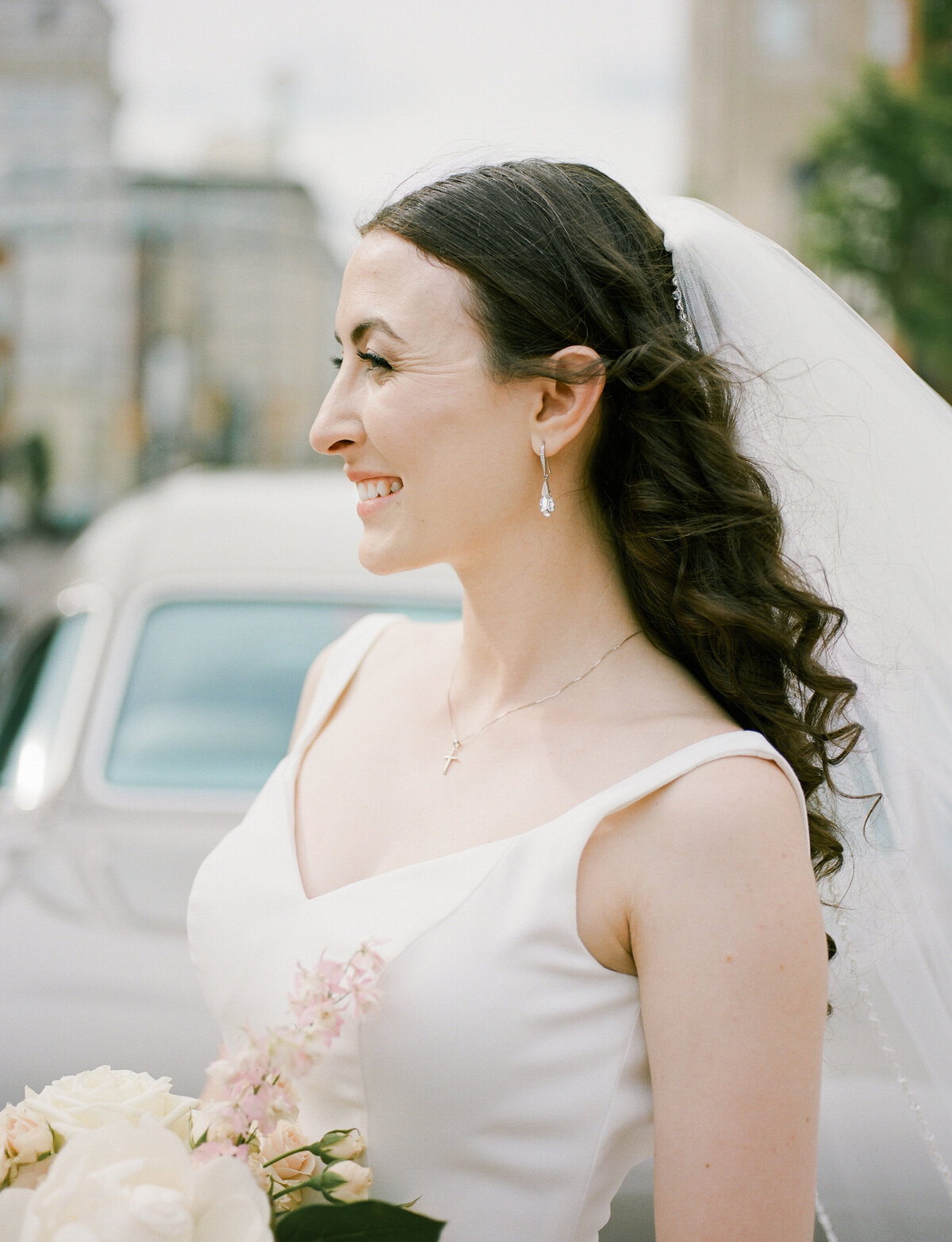 A bride holding her pink and white flowers smiles at her groom in front of a vintage car with the wind blowing her hair and veil gently. Natural makeup by D Neiswender Artistry