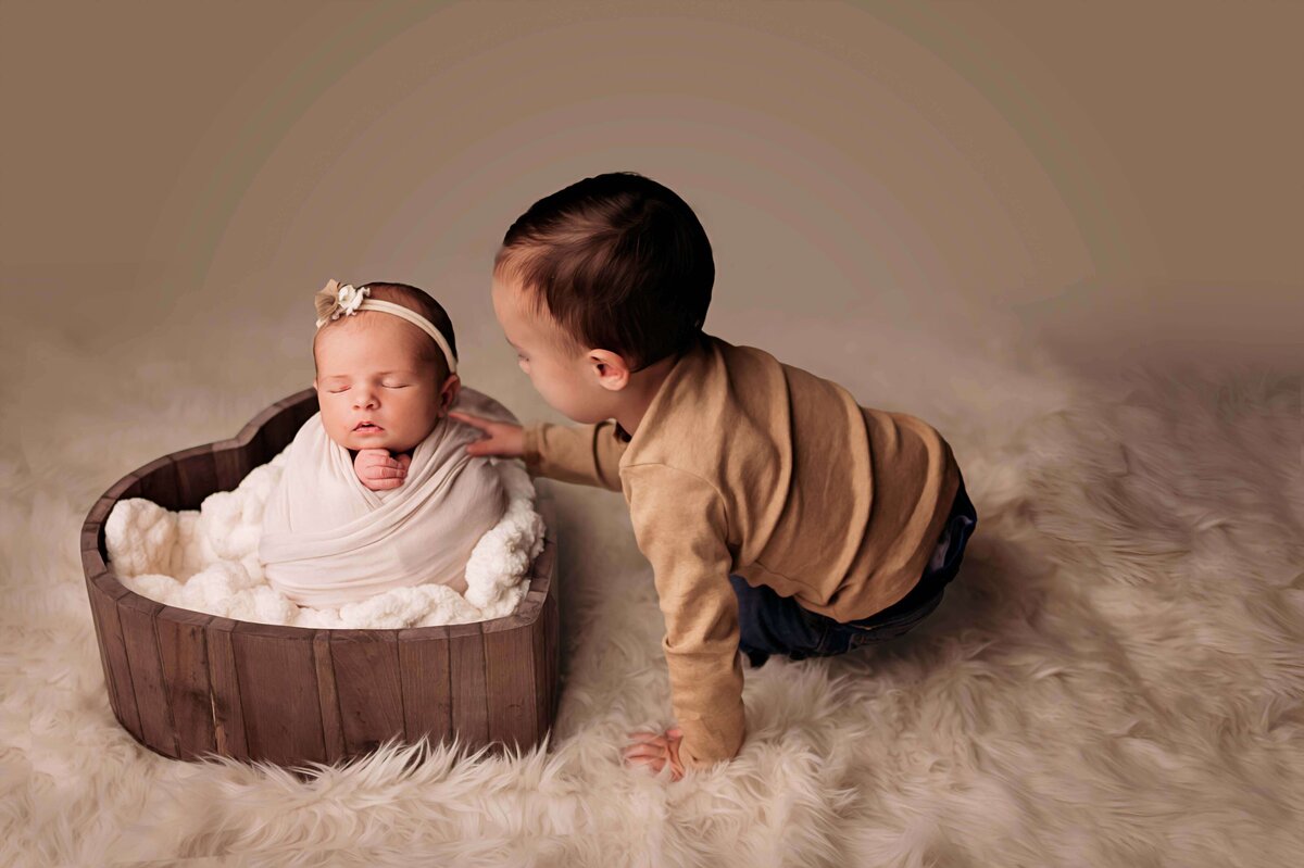 A young toddler boy, checks out his sleeping newborn sister in this perfect newborn photo by Mobile, AL newborn photographer. The boy wears a brown t shirt and blue jeans, sitting a soft cream fur rug, with his newborn sister swaddled in a potato sack inside a heart bowl prop.