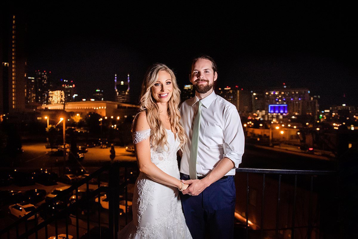 Bride and groom standing on balcony at night with Nashville skyline lit behind them showing iconic buildings