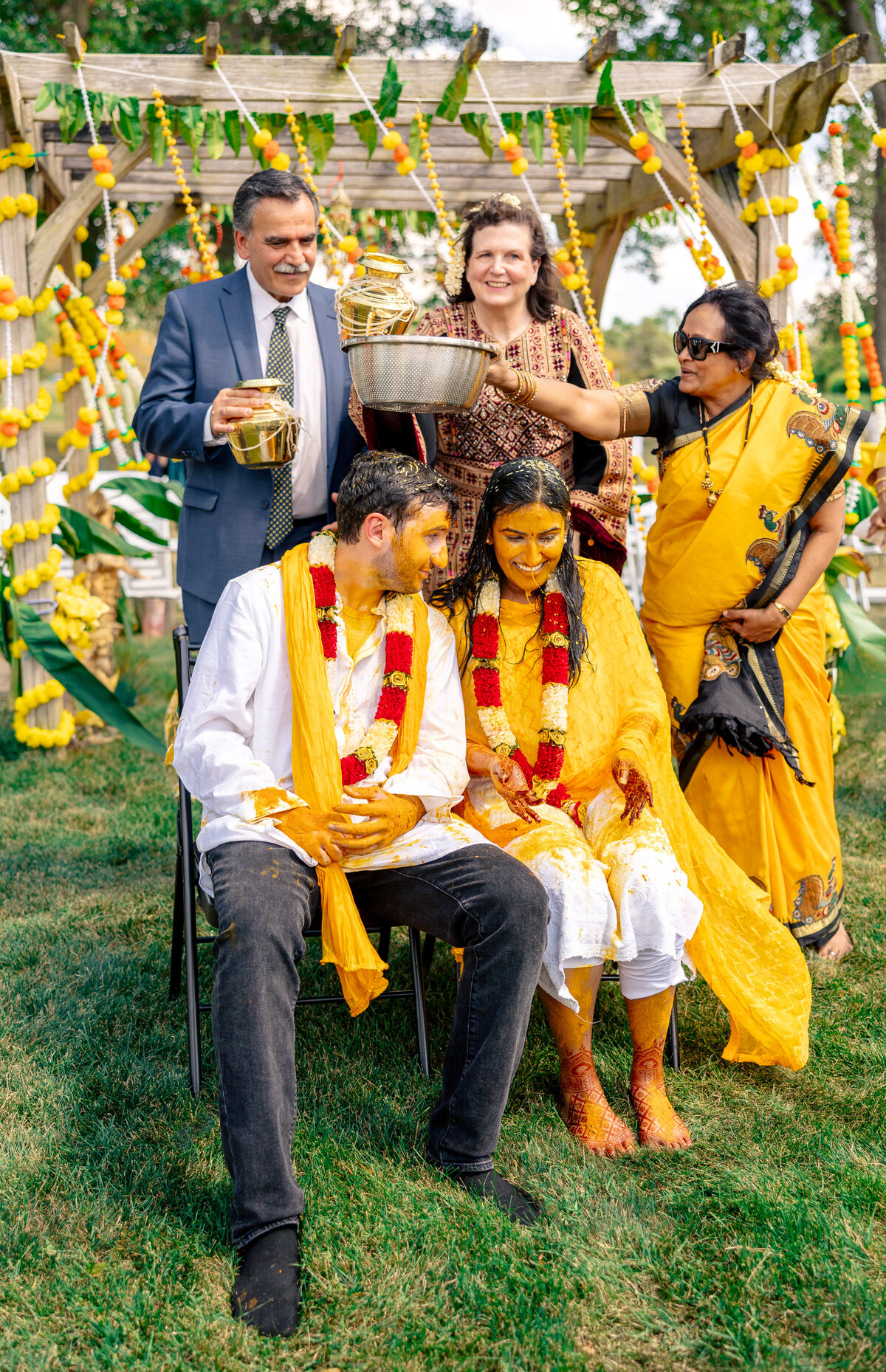A couple covered in turmeric paste sits in chairs during a traditional ceremony. Two women, one in a yellow saree, and a man in a suit pour water over them. The background features colorful flower garlands and greenery.