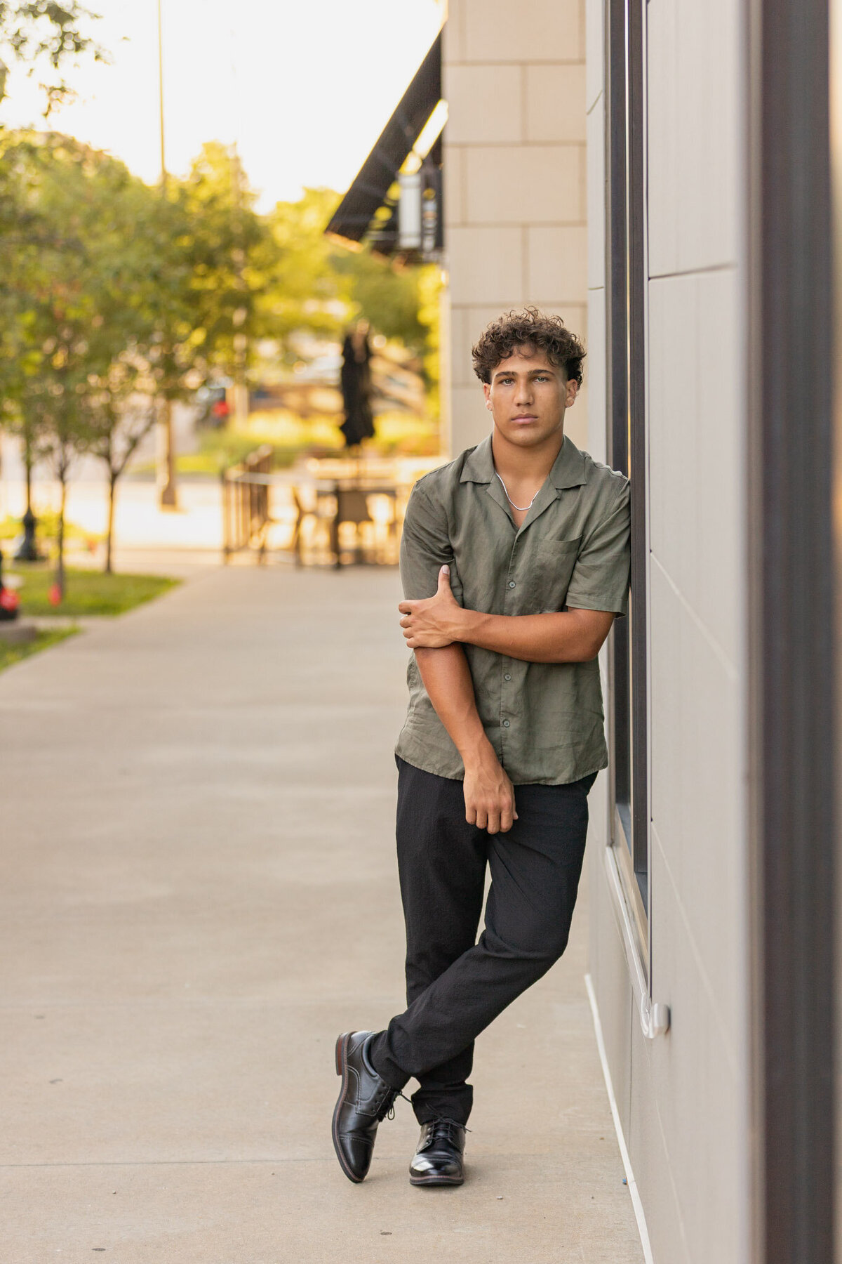 A senior guy leaning up against a wall holding his right arm with his left hand in Lawrence, KS