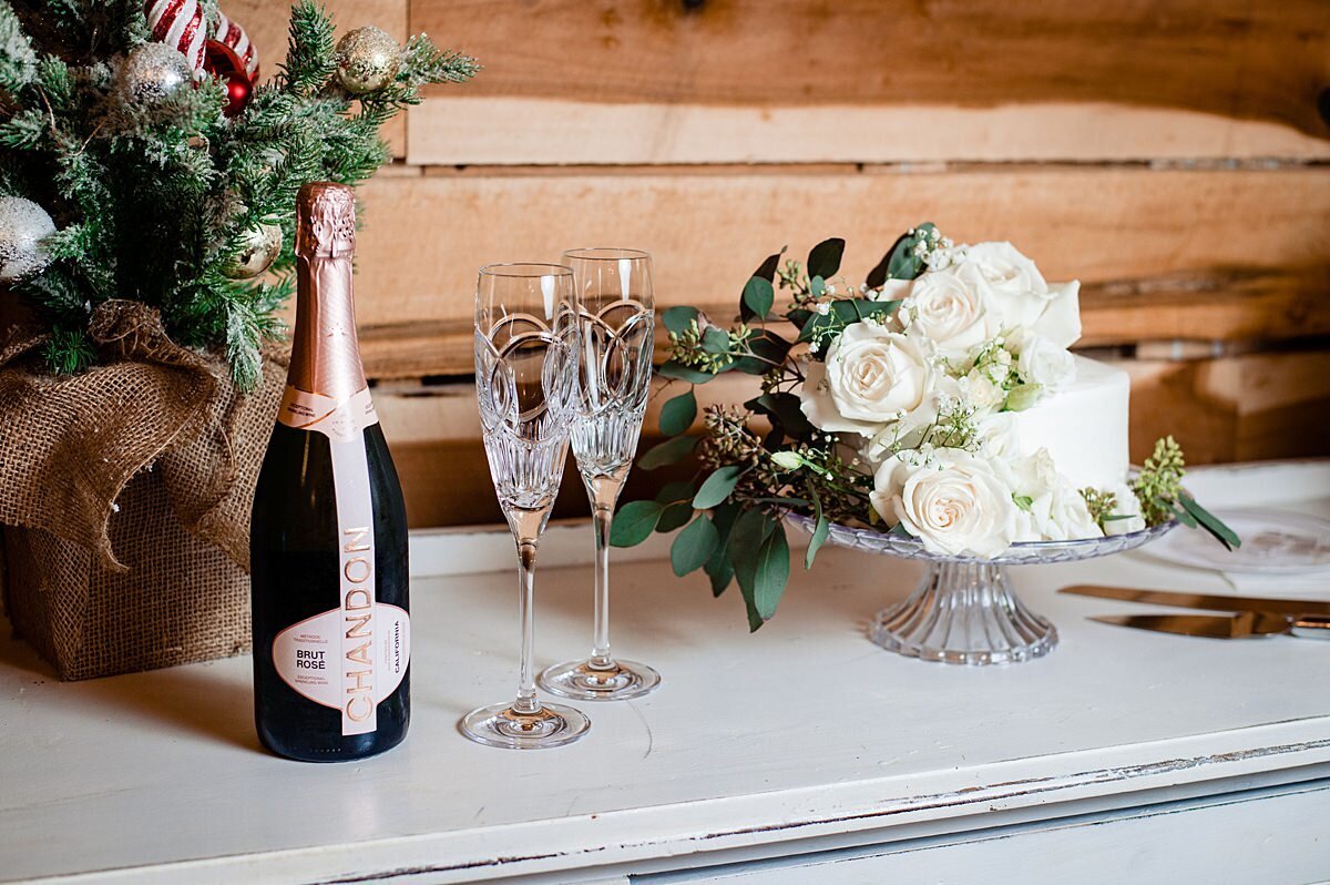 A one tier white wedding cake decorated with white roses and greenery sits on a crystal footed cake stand next to a set of crystal champagne flutes and a green bottle of champagne set on a white farm table with a cedar wood wall behind them. Sitting on the left of the cake is a small Christmas tree for their Christmas Wedding at Steel Magnolia Barn.