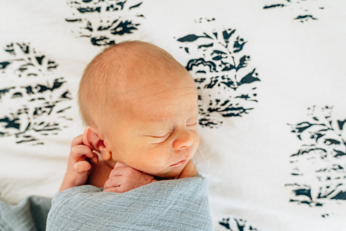 Close-up of a newborn baby wrapped in a blue swaddle, peacefully sleeping on a bed with a black and white patterned bedspread.