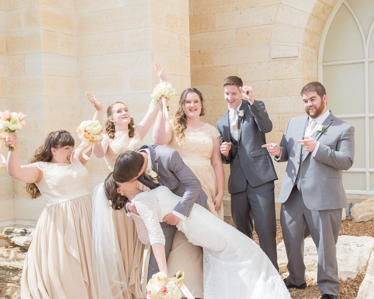 groom dipping and kissing bride in front of a cheering wedding party in cream and gray