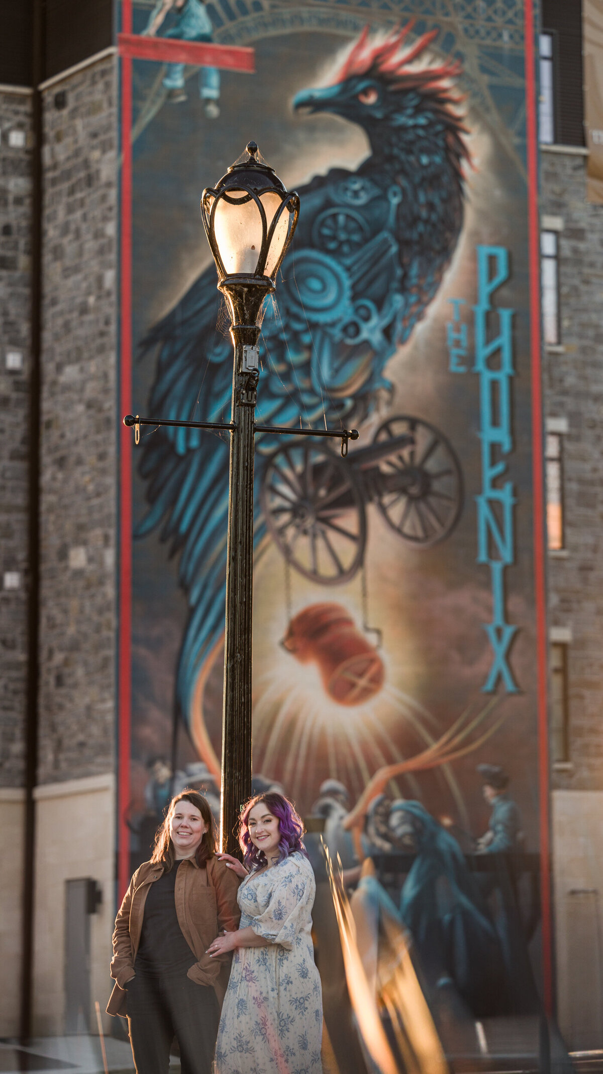 Vibrant engagement photo of a same-sex couple standing in front of a phoenix mural in Phoenixville, celebrating their love amidst a colorful and artistic urban background.