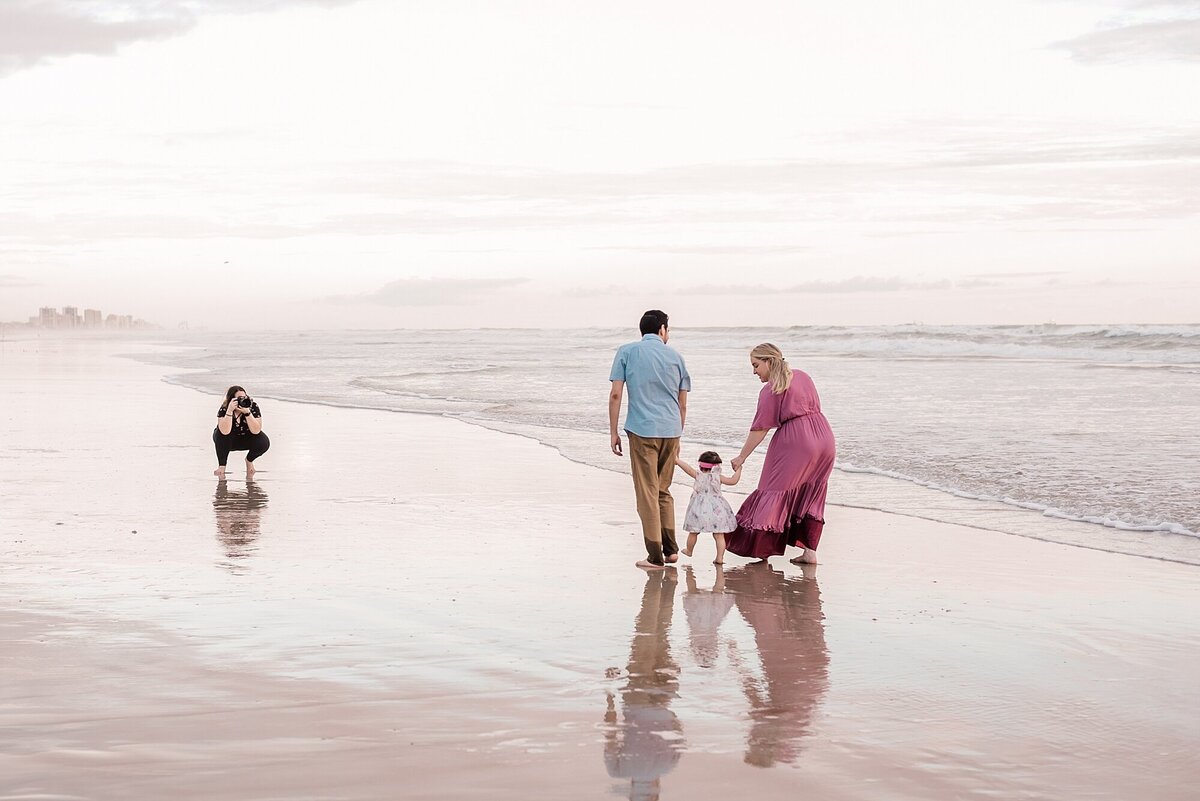 A photographer crouches on the edge of the water at the beach while a couple walks their toddler down to beach towards her