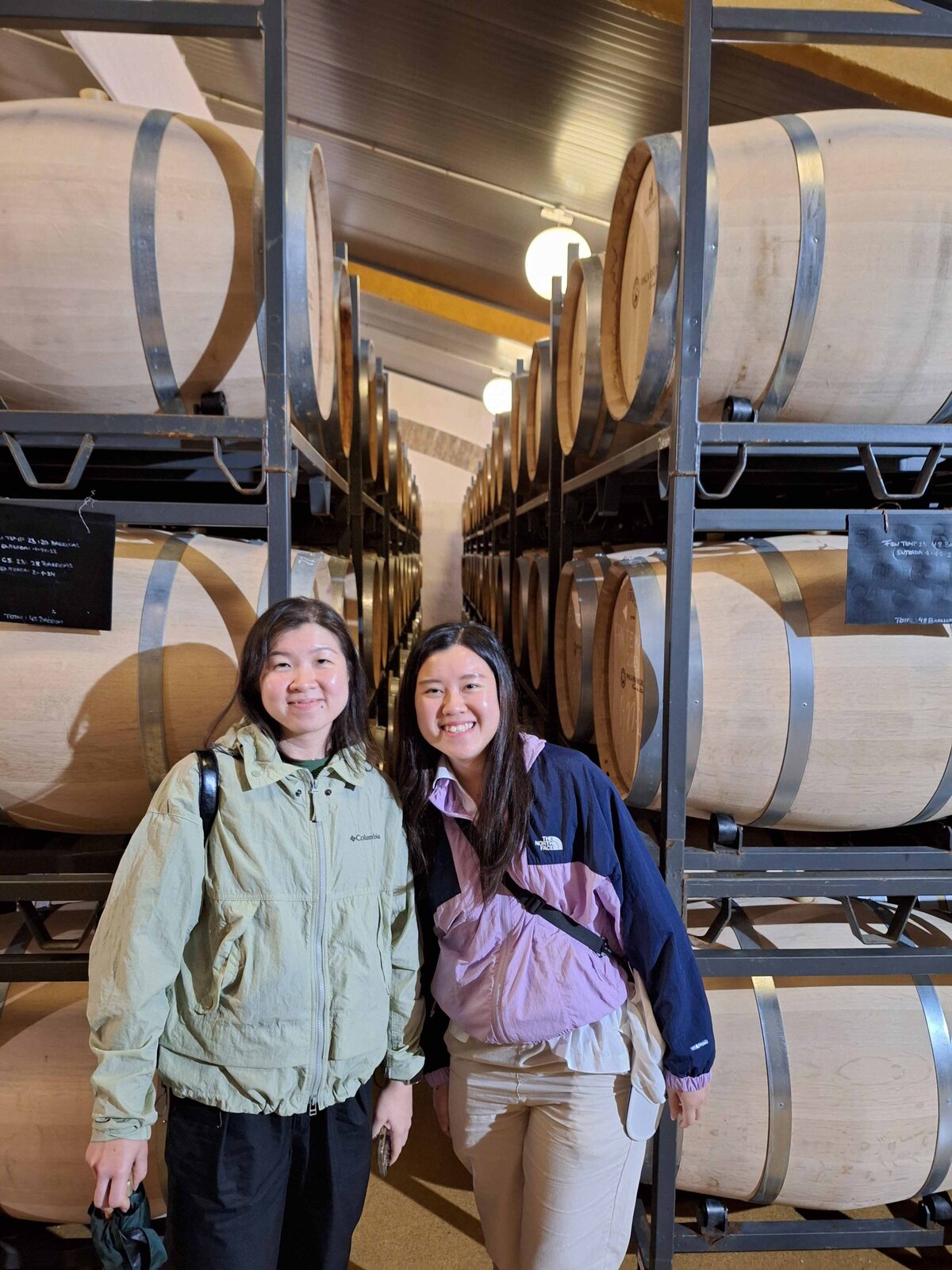 Two women posing for a picture standing in front of wine barrels at the Wine Guide Madrid's Winery Lunch Experience.