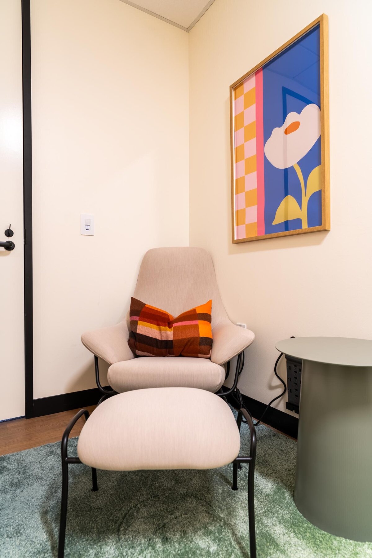 Cozy lounge corner with a cream lounge chair, plaid pillow, ottoman, and colorful floral artwork.