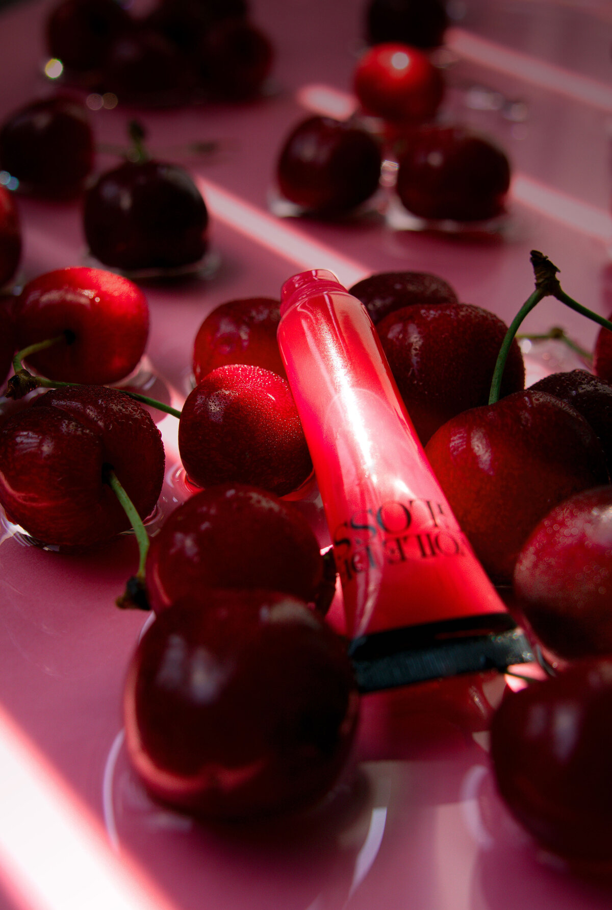 Tube de gloss rouge posé parmi des cerises fraîches sur fond rose, mise en scène gourmande et lumineuse – Studio Helloday.