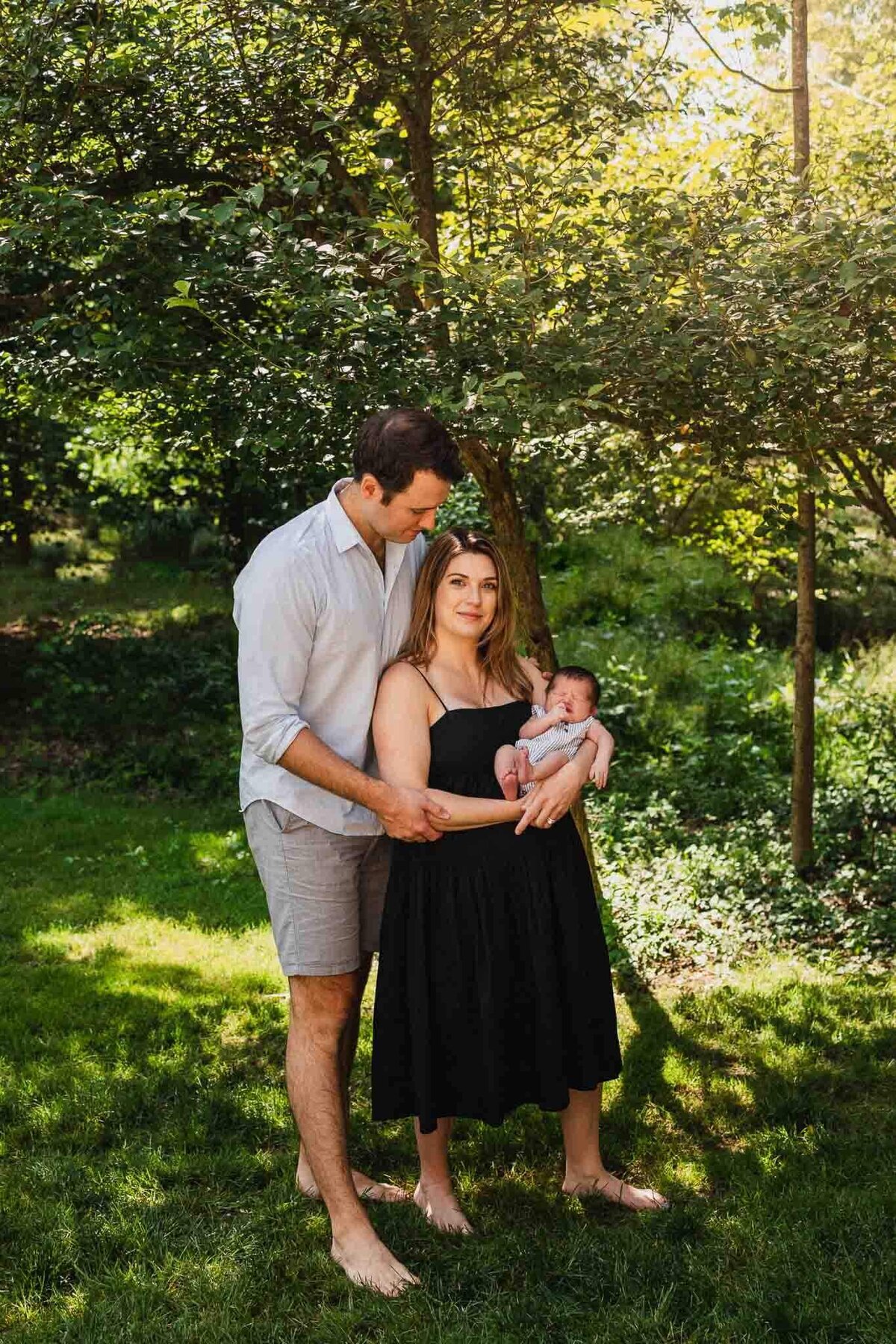 Mom and dad posing with newborn under dappled sunlight is lush green Cleveland yard.