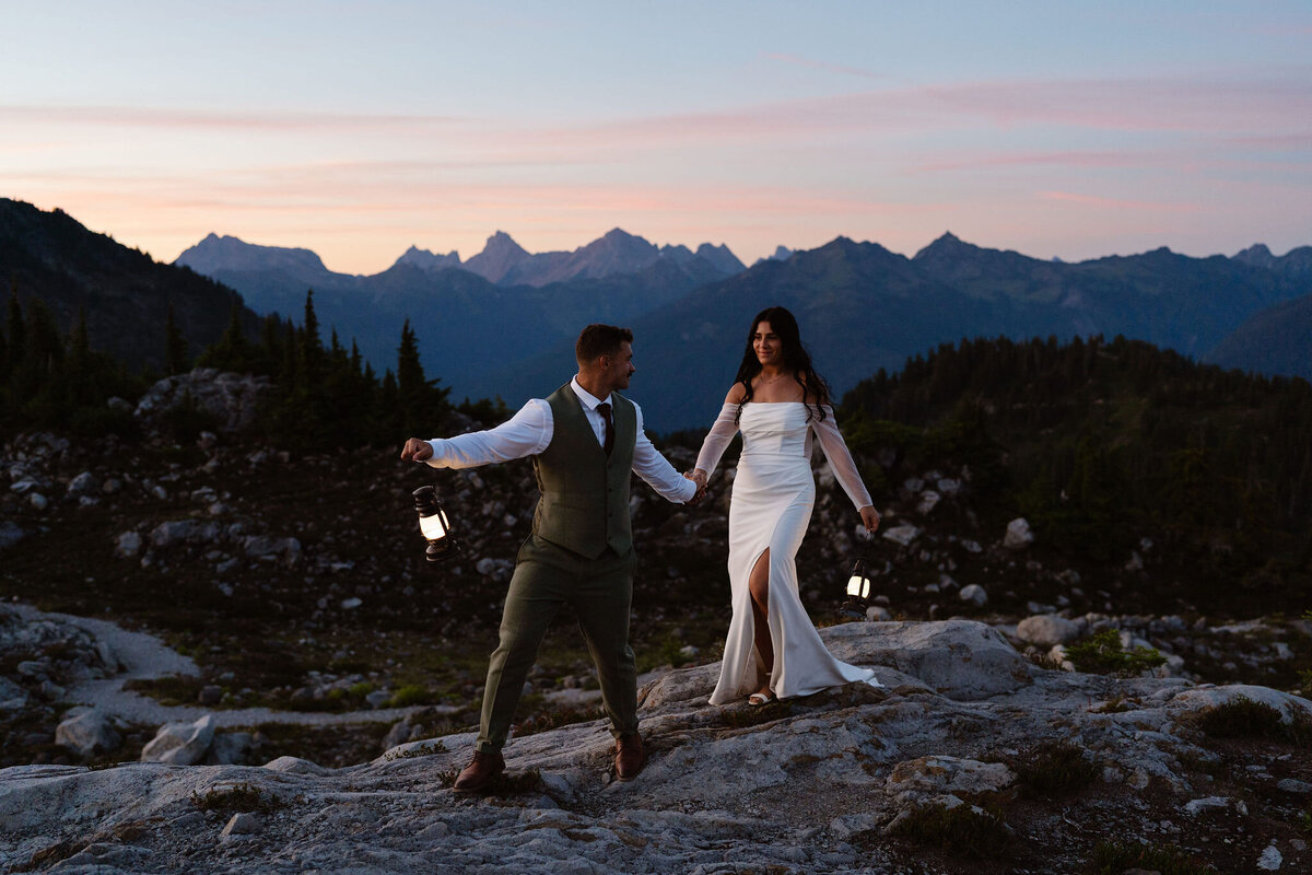 a bride and groom hold hands and lanterns as they walk a ridgeline at sunset during their washington elopement at mount baker